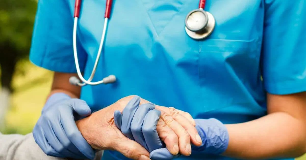 Nurse in blue scrubs and gloves holding an older adult patient's hand for the August Certified Nursing Assistant Course
